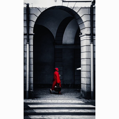 A photograph of a solitary figure in a bright red hooded coat and matching pants, walking through a stone archway. She carries a dark handbag and is wearing  dark shoes. The background features shadowy, arched walls and a textured, cobblestone ground..in the foreground there is a zebra crossing.