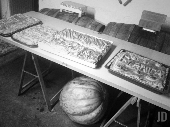 Esta es una fotografía en blanco y negro con un aire rústico y tradicional. Esto es lo que se observa:
• Primer plano: Debajo de una mesa sostenida por caballetes, destaca una calabaza de gran tamaño.
• Sobre la mesa: Se muestran varias bandejas con hornazos típicos de Salamanca. Una de ellas tiene el nombre "BLANCA" moldeado en la masa.
• Fondo: Se aprecian varios barriles de madera alineados contra la pared y una pequeña caja de madera (con el texto "Fresón de Palos") encima de ellos.
La escena sugiere una preparación para una celebración local o familiar, posiblemente en una bodega o un espacio rural.