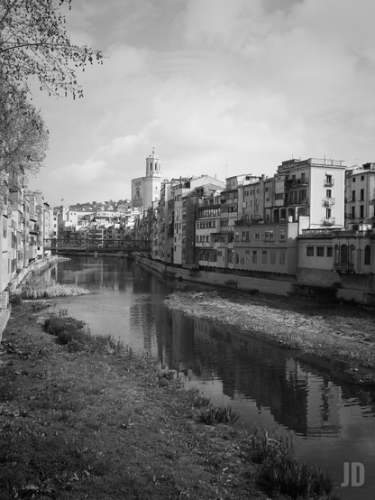 Esta es una evocadora fotografía en blanco y negro de Girona, España. La imagen captura una vista clásica de la ciudad con los siguientes elementos destacados:
• Las Casas del Onyar: Los edificios icónicos que bordean el río, cuyas fachadas se reflejan suavemente en el agua.
• La Catedral de Girona: Al fondo, se alza majestuosa la torre de la catedral, dominando el horizonte urbano.
• El Puente de Hierro: Se alcanza a ver el famoso puente (Pont de les Peixateries Velles), diseñado por la compañía de Gustave Eiffel.
• Atmósfera: El uso del blanco y negro resalta las texturas de la piedra, el curso tranquilo del río y las nubes en el cielo, dándole un aire atemporal y nostálgico.