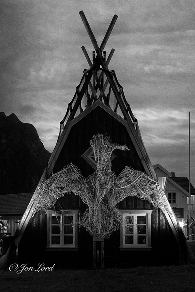 This is a night-time photo of the gable end of a Norwegian summer cabin made from the frame of a fish drying frame (Hjell) with a number of fishing nets arranged on the wall into the shape of a large bird. Svolvær, Lofoten Islands, Norway (2018).

Rising from the lower left and right corners is a wooden frame forming an inverted 'V' or Isosceles triangle, viewed end-on, with the shorter length being on the ground. The frame stretches away from the camera forming a wedge shape (think Toblerone chocolate box shape). From the end of the frame are long horizontal poles that are spaced at one metre intervals up the height of the frame and run from one end to the other end. Draped over the frame is a large fishing net or nets to deter birds from feeding on the drying fish hanging from the frame sometimes called a flake, though the fish are absent here. Within the frame is a small dark wooden building shaped much like a simple 'Vee' shaped tent - all roof and no walls except at the ends. Here at the viewers end are two ground level, white, rectangular windows with a single square window above. Two floodlight illuminates a further set of fishing nets that have been arranged into the shape of a large bird or possibly bat with the wings outspread . Above and to the upper left of our building is the silhouette of a distant mountain descending from left to the right. Above is a thin overcast sky with almost translucent Cirrus clouds dimly lit by the near midnight sun.