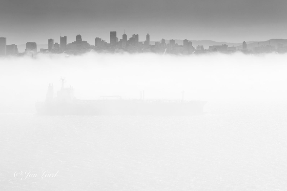This is a black and white photo in landscape format of a ship underway through a mass of thick sea fog with a cityscape in the background. San Francisco, California (2016).

The mass of fog fills the photo from the base upwards to a little over the three-quarters level. In the middle of the image and the fog, is a large cargo ship (crude oil tanker) underway from left to right, viewed in profile and filling about two-thirds of the length of the photo. The ship is travelling through the sea fog and is only partly visible, the hull is dark and the wheelhouse / accommodation superstructure is at the stern. The top of the fog bank is rough and wispy. Stretching along the top of the fog and in the middle ground from left to right is a high-rise cityscape a few km away. In the background and the upper right corner are a few distant hills. Above is a clear and cloudless sky. 