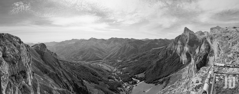 Esta es una impresionante fotografía panorámica en blanco y negro que captura un vasto paisaje montañoso desde un punto de observación elevado.
Aquí tienes los detalles principales:
• Composición: La imagen muestra un valle profundo y serpenteante en el centro, rodeado por picos escarpados y laderas densamente arboladas. Se puede apreciar una pequeña carretera o sendero que recorre el fondo del valle.
• Primer plano: A la derecha, se observa parte de una barandilla metálica con pegatinas, lo que sugiere que la foto fue tomada desde el mirador de la Estación Superior del Teleférico de Fuente Dé, en los Picos de Europa. 
• Geología: Destacan las formaciones rocosas calizas, con paredes verticales y crestas afiladas que contrastan con las zonas de vegetación más oscura.
• Atmósfera: El uso del blanco y negro resalta las texturas de la roca y el juego de luces y sombras bajo un cielo parcialmente cubierto con nubes ligeras.