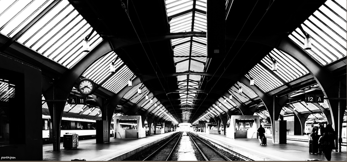 A black-and-white photo of a platform with a glass roof, a visible clock, benches, and a few people waiting near tracks 11 and 12.

Schwarz-Weiss-Foto eines Bahnsteigs mit Glasdach, einer sichtbaren Uhr, Baenken und einigen wartenden Personen in der Naehe der Gleise 11 und 12.