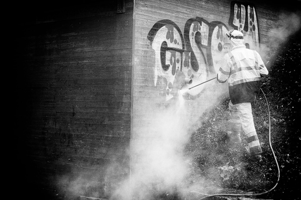 A man in protective work clothes is removing a large graffiti by a bridge tunnel entrance with water.