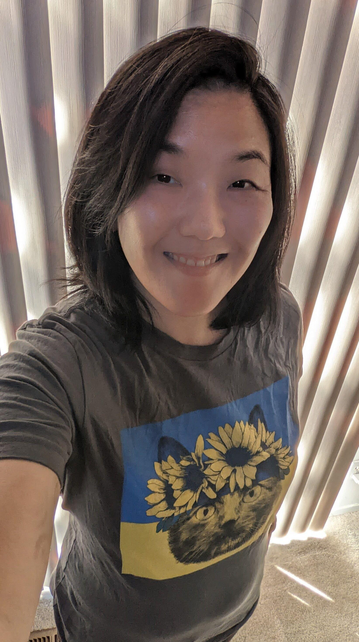 Photo of an Asian woman with medium long hair wearing a gray tshirt with a painting of a cat. The cat is wearing a crown of yellow sunflowers, and the background is the blue and yellow of the Ukrainian flag. 