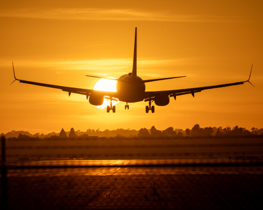 Looking down an airport runway at sunset, with the silhouette of an arriving Southwest Airlines Boeing 737 overlapping the sun. The airport fence is visible in the foregound, and the sky is golden.