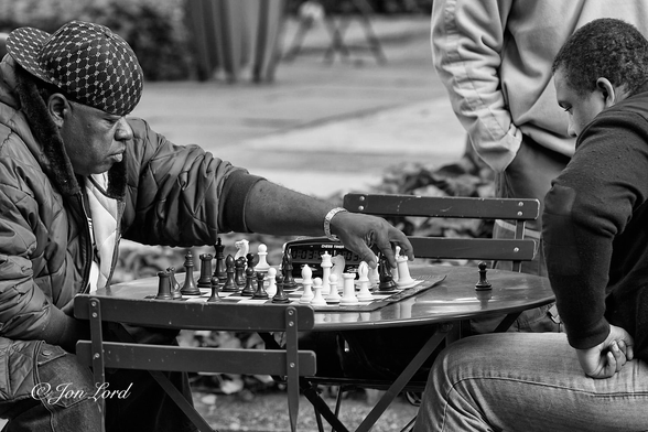 This is a black and white street photo in landscape format of two men playing chess. Bryant Park, NYC (2012).

Sitting on a park chair on the left side of the photo is a young African-American man, viewed in profile, wearing a dark patterned baseball cap, turned backwards to the rear, a dark, thick anorak and having a serious expression on his face. His left arm is extended as he moves a chess piece over to the far side of the chessboard that sits on a small, dark, round metal table. Sitting opposite is a further African-American man. He has short dark hair, is wearing a dark winter's jacket and has his left fist clenched on his left thigh. He to is staring intently at the chessboard. Between these two men and at right angles are two empty chairs. On the far side of the table is a digital chess clock. A person is watching the game and is partially visible in the upper right corner. 