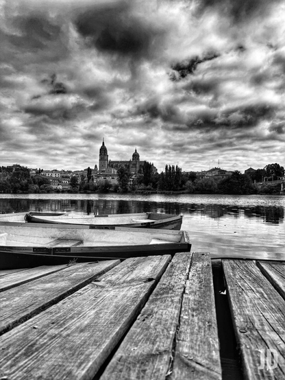 Esta es una fotografía artística en blanco y negro con un alto contraste que captura una vista serena y algo dramática de la ciudad de Salamanca, en España.
La composición se divide en varios planos interesantes:
• Primer plano: Un muelle de madera con texturas muy marcadas que guía la mirada hacia el agua.
• Plano medio: Dos pequeñas barcas amarradas en el río Tormes, que reflejan la luz del cielo.
• Fondo: El perfil de la ciudad, donde destacan las icónicas torres de la Catedral de Salamanca y la cúpula de la Clerecía, rodeadas de vegetación.
• El cielo: Un manto de nubes densas y oscuras que le otorga a la imagen una atmósfera imponente y melancólica.
