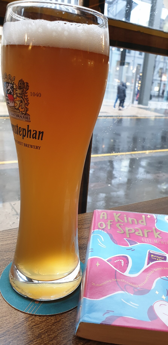 A window seat in a British pub. Outside, it is miserable, windy and pissing it down. But inside, there is a tall pint of wheat beer, and a book ('A Kind Of Spark' by #ElleMcnicoll) with  a cheerful cover of pink fuchsia waves.