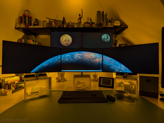 A five-monitor computer setup bathed in a warm, amber-toned light. The workspace is organized but packed with personal items.

Three large monitors are aligned horizontally in the foreground. They display a unified panoramic wallpaper of a planet's curved horizon against the blackness of space. Mounted above the center and right foreground monitors are two smaller screens, each featuring a different celestial body.

The background is dominated by a long shelf filled with a dense collection of books and collectibles. Two small stuffed toys are tucked underneath the monitors on the desk surface.