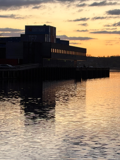 Orangegelber Himmel mit anthazitfarbenen Wolkenstreifen über dem Ostseewasser des Alten Hafens in Wismar, in dem sich das Orange dew Hinmels spiegelt. Links im Bild die schwarze Silhouette eines Hafengebäudes, in dessen Fensterreihe sich ebenfalls das Orange des Sonnenuntergangslicht spiegelt. Das dunkle Hafen-Gebäude spiegelt sich im Meerwasser.  