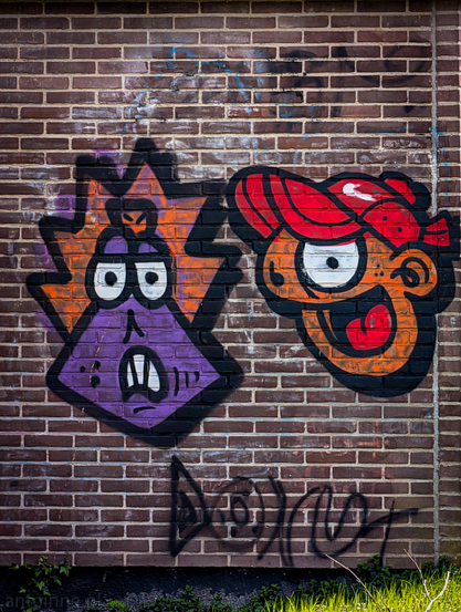 A vibrant piece of street art painted onto a brick wall. The graffiti depicts two stylized, cartoonish characters side-by-side, each with a distinct personality and color palette.

Both figures are defined by thick, bold black outlines, a hallmark of classic graffiti "character" styles. Below the main characters, there is a faint, dark "tag"that appears to read "DONUT".

At the very bottom of the frame, small green plants and blades of grass peek up from the base of the wall, contrasting the industrial feel of the brick with a touch of nature.