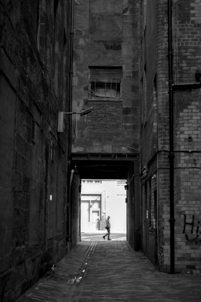 Black and white street photo taken from a dimly-lit alleyway between tall buildings and looking out to a brightly-lit main street in which a passer-by is seen walking across the entrance to the alley and glancing toward the camera.