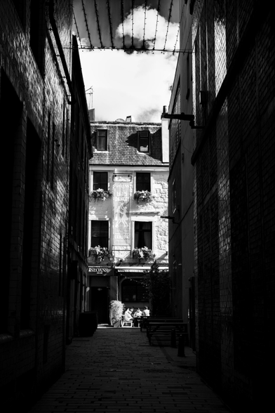 Black and white scene captured in a dark alleyway between tall buildings in which a group of people sit in the sunshine outside a bar. These modern, urban 'canyons' often providing striking patches of light and shadow.
