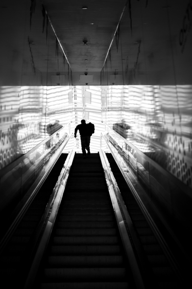 A man ascends a moving staircase into a shopping centre, going from the dark, shadows into bright light where he is just a silhouette 