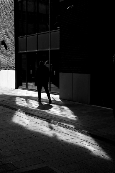 Patches of reflected sunlight from glass-fronted buildings fall onto the ground in a narrow alleyway and a man dressed in black is caught in silhouette as he walks through the scene