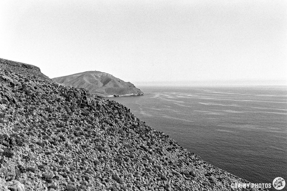 A monochrome photograph showcasing a rugged coastline with rocky cliffs leading down to a calm sea. A distant mountainous area is visible in the background, creating a serene, natural landscape.
