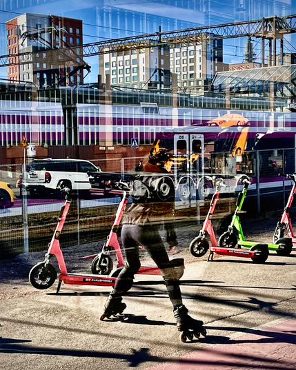 A photographs of a person rollerblading near a row of electric scooters in front of a train station, with buildings in the background. Reflections of cars from a bus shelter glass wall can be seen too.