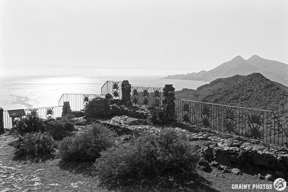 Scenic view from a rocky vantage point overlooking a sparkling ocean. A rustic stone wall and railing frame the landscape, with hills and mountains visible in the distance under a clear sky, creating a serene and picturesque atmosphere.