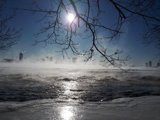 Looking across a wide, mostly frozen over river with some rapids near the foreground keeping the water open. Across the river there are the silhouettes of buildings through the heavy evaporation coming off of the water into the bright sun behind.