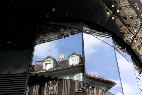 Ein Hausdach mit zwei Dachfenstern und einer alten Satellitenschüssel spiegeln sich in einer Glasfront eines modernen Gebäudes.