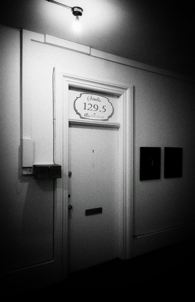 A closed white interior door with a sign reading "Studio 129.4" above it is seen in this black and white image. A bare light bulb hangs from the ceiling lighting the door. Black boxes are attached to the walls and a mail slot is shown on the door.