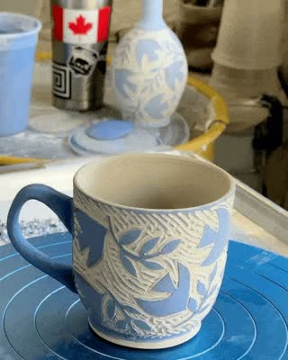 a gif image of a work in progress pottery cup rotating on a wheel. it is decorated with blue doves and olive branches.