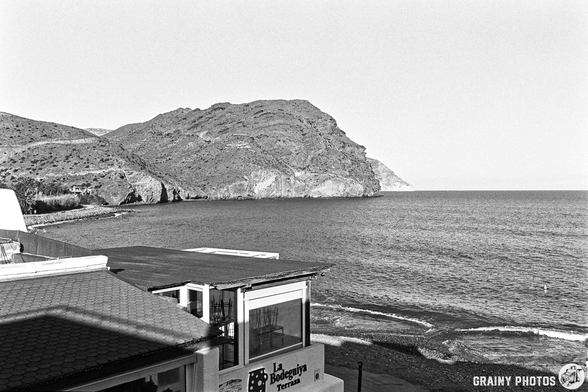 A monochrome view of a coastal landscape featuring a rocky hill, a calm sea, and a building with the name "La Bodeguiya." The scene captures a tranquil atmosphere with slight waves and a distant silhouette of the mountain.
