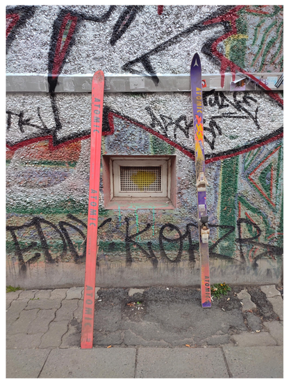 Farbfoto im Hochformat mit weißem Rand. Zwei alte bunte Skier stehen an einer mit Graffiti bedeckten Hauswand.
