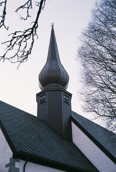 Photograph showing the tower and roof of a medieval stone church, modernised throughout the centuries. The walls are painted white, the roof is black and the wooden tower a dark brown with a matching spire on top. The scene is framed by the naked branches of surrounding trees against a clear but pale sky. It's a colour photo, but you'd be forgiven for thinking it was monochrome.