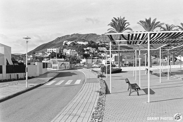 A quiet street scene in black and white, featuring palm trees, a curving road, and a pedestrian walkway. In the background, a hillside town with white buildings is visible. Modern architecture and shaded seating areas create a serene atmosphere.