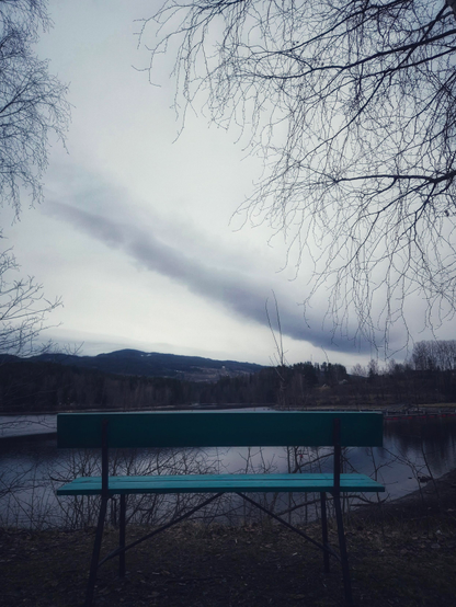 A green bench in front of a lake and rolling hills on the background. Only one strange dark long cloud sets a contrast to the otherwise very white sky. The picture is quite dark, since it was taken towards the light sky.