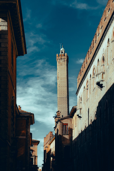 This is a photograph down a  narrow, sunlit street (via Castiglione) which leads towards a tall, slender medieval tower. (Asinelli Tower in Bologna). On either side of the street are historic multi-story buildings with textured masonry, arched windows, and some small stone balconies. There are shadows which stretch across the lower portions of the buildings. The sky above is a vibrant deep blu and there are wispy, streaking white clouds.