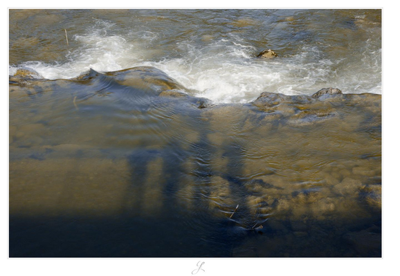 From a bridge over a river we are looking down to the water that flows over a ledge in the upper third of the picture and forms white foam and vortexes. On the surface of the calmly floating water below, the shadow of the bridge and four people leaning against the railing can be seen, casting from the sun behind. The colors of the river are brownish-green and white.

AI disclaimer: Using my work, its meta data, written or derived description to create media with or train AI based systems is prohibited.