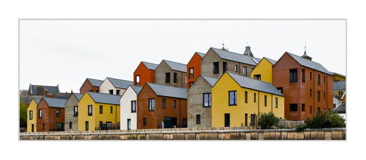 Wide horizontal color photograph. A row of modern houses fills the lower half of the image, arranged in overlapping layers along a waterfront. The buildings have simple gabled roofs and façades in muted yellow, orange, red brick, cream, and pale gray brick, with dark window openings and slate-colored roofs. In the foreground, a low wall runs horizontally across the bottom edge. The upper half is mostly a pale, overcast sky, while a few older rooftops and small tower-like structures are visible behind the newer houses.