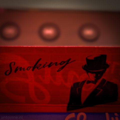 The image is a stylized, close-up photograph featuring a red rectangular surface with vintage-style graphic design. The word "Smoking" is written in a black, elegant script font across the center-left. On the right side, there is a high-contrast illustration of a man in a tuxedo and top hat. His face is mostly obscured by shadow, giving him a mysterious or "gentlemanly" appearance typical of early 20th-century branding.