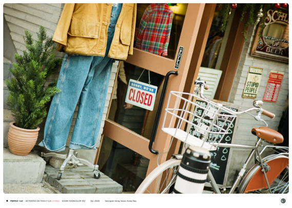 열린 가게의 나무 문 옆으로 탁한 노란색의 외투와 청바지를 걸친 마네킹이, 그 앞으로는 레트로한 스타일의 자전거가 주차되어 있는 모습.