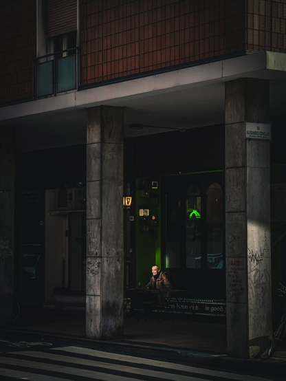 A photograph of a dimly lit urban street corner at late afternoon. A person with short hair and a beard who is wearing a brown jacket is sitting at a small pub table under a concrete overhang. Their face is illuminated by a sunbeam reflected off something (I think it's a car but I didn't find what it was!) The building has a tiled facade and graffiti on the pillars. A green lamp glows in the background inside the pub. There is a zebra crossing is in the foreground on the road.