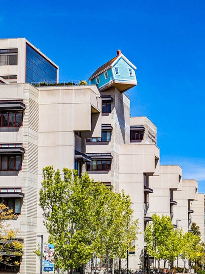 An art installation of a small, blue New England-style cottage titled "Fallen Star" perched precariously at a tilted angle on the edge of the roof of Jacobs Hall, a modern concrete engineering building at UC San Diego.