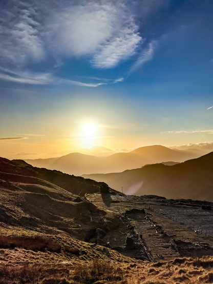 A landscape shot of the Moelwynion mountains in Eryri, North Wales at sunset. The foreground shows the stone ruins of a slate quarry on a grassy hillside. The setting sun is low on the horizon casting a golden glow over the distant peaks and a valley filled with mist.