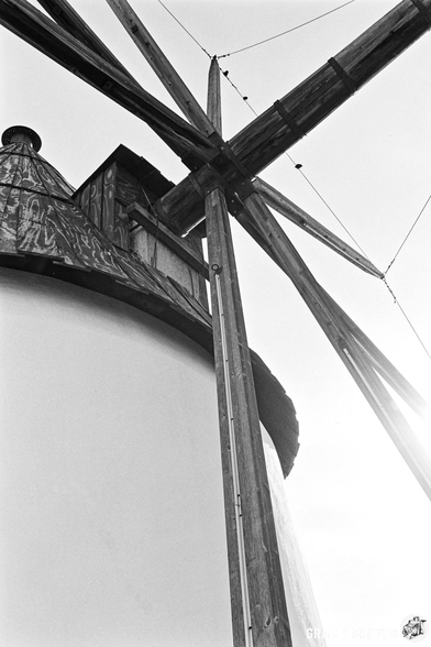 A close-up view of the wooden structure of a windmill, featuring its large crossarms and circular base. The image is captured in black and white, highlighting the textures of the wood and metal components against a soft sky.
