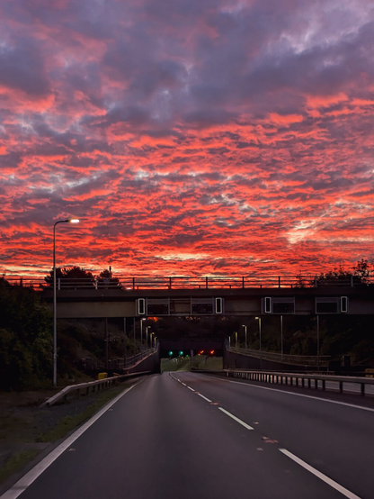 A scenic view of a highway at dusk under a breathtaking "fire sky." The sky is full of vibrant, glowing red clouds that stretch across the entire horizon. Underneath, the dark road and the silhouette of a bridge are in contrast.