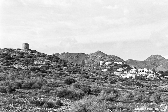 A black and white landscape featuring rolling hills, a prominent abandoned windmill on a hill, and the village of Las Negras with white buildings nestled among the mountains under a cloudy sky.