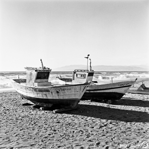 Two weathered fishing boats resting on a sandy beach, with waves crashing in the background and a clear sky. The scene captures the calm yet rugged beauty of coastal life.