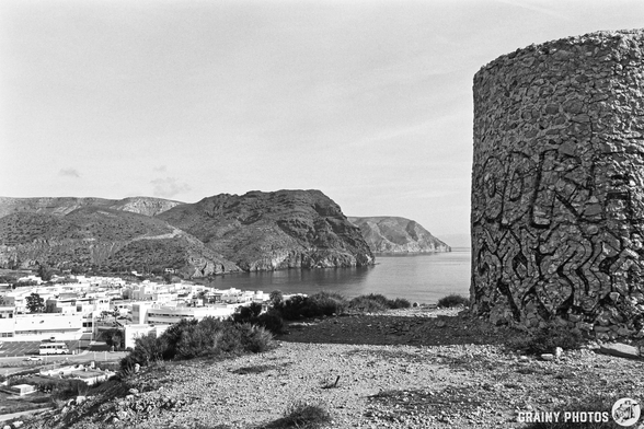 A rocky coastal landscape featuring a weathered ruins of a stone windmill adorned with graffiti, overlooking las Negras on the coast, with distant hills and a cloudy sky above.