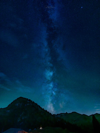 A stunning night sky filled with stars and the Milky Way, positioned above dark mountain silhouettes. A small structure is illuminated in the foreground, enhancing the tranquil atmosphere.