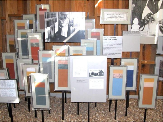 A display of rectangular paint swatches displayed vertically on small black stands. Each swatch is divided in three sections with two shorter stripes on top and a third, larger block of color on the bottom. The swatches were meant to suggest different color combinations for ceiling, trim and walls. The tones are earthy and pastel, from pink and light blue to brick red, orange and yellow ochre. There are small accompanying signs sharing information about George Washington Carver and the title of this book "White and Color Washing with Natural Clay from Macon County, Alabama."
