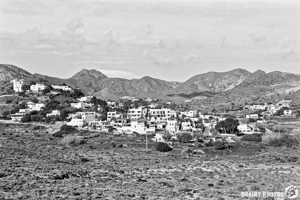 A black and white landscape featuring a Las Negras nestled in a valley. The scene is surrounded by rolling hills and mountains, with scattered homes and vegetation visible, evoking a serene and rustic atmosphere.