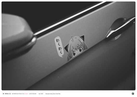 흑백 사진. 한 차량의 운전석 도어에 '아냐 포저' 캐릭터가 힐끗 쳐다보며 'わくわく'라 말하는 스티커가 붙여진 모습.