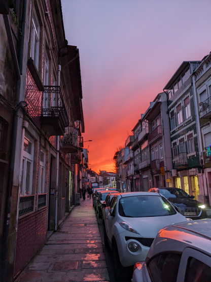 A Braga, Portugal street at sunset lined by low rise buildings. Balconies alternate almost every building on the second floor. The one way street is narrow and lined with cars parked to the left. The concrete paved sidewalk has people walking home from work down them.  The sky blazes orange-red with clouds in the far distance over a closer purple-blue sky above. Reflections of the sky are seen from the cars.
